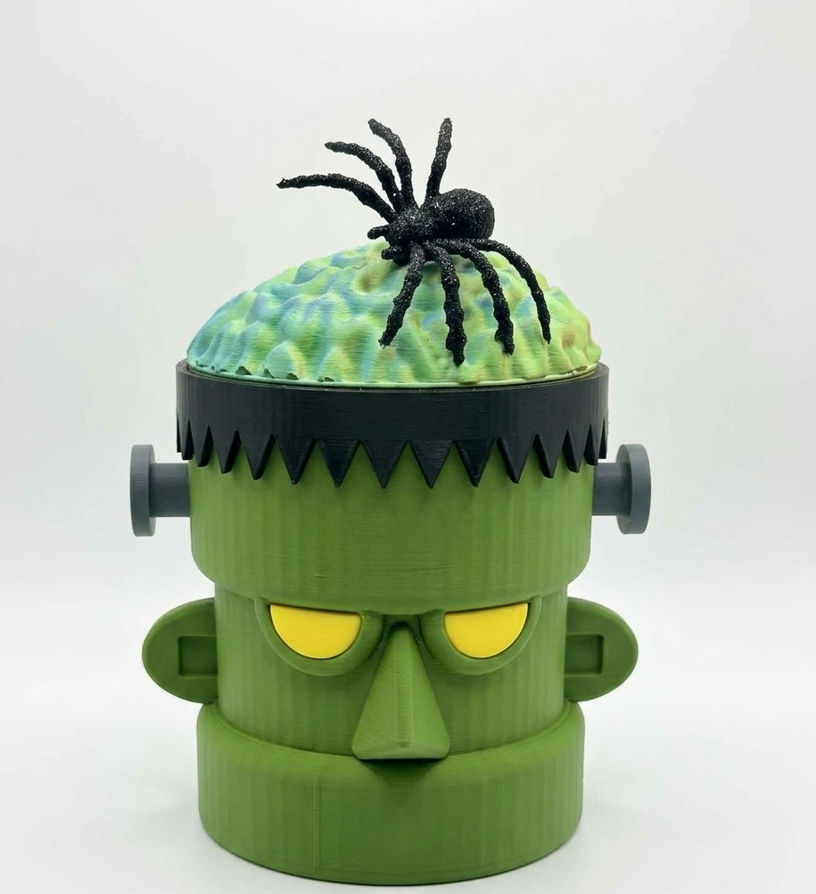 A 3D-printed Frankenstein's monster head container or decor piece in green, with yellow eyes, a black hair band, gray side bolts, a green brain-like top, and a black spider on top, photographed against a plain white studio background.