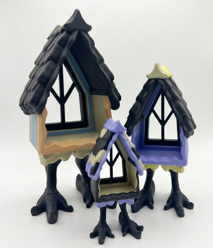 Three small decorative treehouse-style figurines with black clawed legs and pointed roofs, shown on a plain light background. The left house is beige and black with blue trim, the right house is purple and black with a pale yellow roof peak, and the center house is beige and purple with black trim.