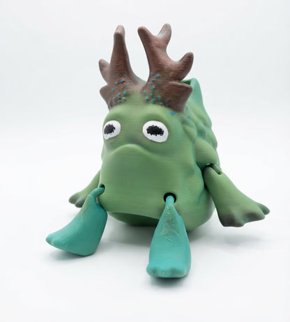 A green cartoon monster toy with brown antler-like horns, large white eyes, and teal legs, photographed on a clean white studio background.